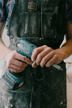 Crop anonymous electrician in uniform with wireless drill standing in flat during repair works